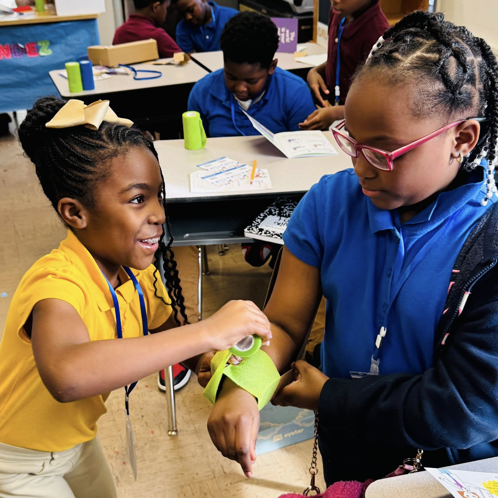 A little girl wrapping green gauze around another little girl's arm.